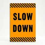Slow Down sign with black text on orange background and diagonal black and orange hazard stripes