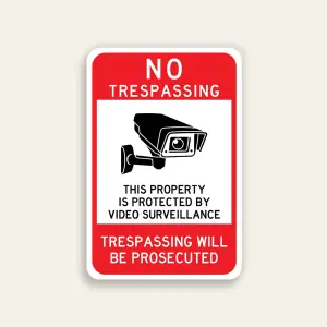Full red 12x18 vertical Maxmetal sign with bold white NO TRESPASSING in upper red zone, large white inset panel with oversized front-facing CCTV camera illustration and black text This Property Is Protected By Video Surveillance, and bold white TRESPASSING WILL BE PROSECUTED on red lower panel.